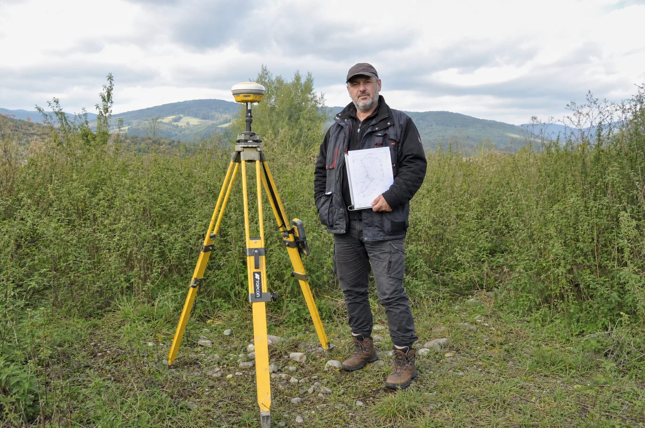 Dokumentárna foto: muž v pracovnom outfite kontroluje pozemok s papierovým plánom v ruke, vedľa neho geodetický GPS prijímač na trojnožke, zarastená parcela v slovenskom podhorí.